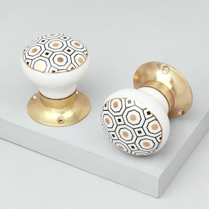 Op de afbeelding: Twee witte keramische deurknoppen met een geometrisch patroon in zwart en goud. De knoppen zijn gemonteerd op messing platen.