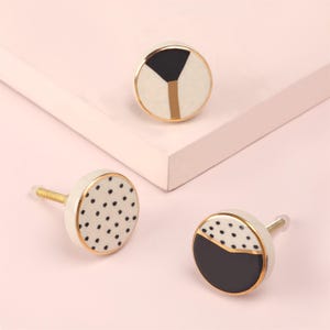Könnte beinhalten: Drei dekorative Schrankknöpfe mit goldenen Akzenten. Ein Knopf hat ein schwarz-goldenes geometrisches Design, während die anderen beiden eine weiße Basis mit schwarzen Punkten und ein schwarz-weißes Halbkreis-Design haben.