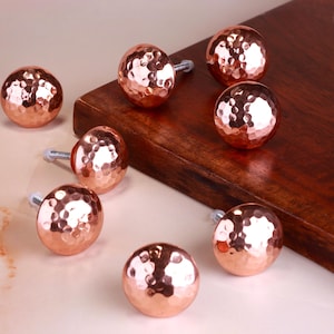 May include: A collection of eight copper-colored, hammered-finish cabinet knobs. Each knob is spherical with a textured surface and a silver-colored screw for attachment. The knobs are arranged on a light-colored surface and a dark wooden board.