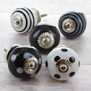 Puede incluir: Un conjunto de cinco pomos decorativos de cerámica para armarios. Los pomos presentan varios diseños, incluyendo rayas blancas y negras, lunares y negro liso. Cada pomo tiene una base y un tornillo de metal plateado para la instalación.