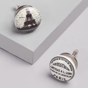 Peut inclure: Deux boutons de meuble décoratifs. L'un présente une image en noir et blanc de la Tour Eiffel avec une écriture. L'autre bouton porte l'inscription "Fournitures de pierres de villebois, POUN SOCLES ET BALCONS, PARIS."