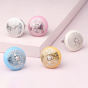 Könnte beinhalten: Eine Sammlung dekorativer Schrankknöpfe in verschiedenen Farben: Weiß, Rosa, Blau und Gelb. Jeder Knopf hat ein silberfarbenes, durchbrochenes Design über einem soliden Sockel. Die Knöpfe sind rund und haben eine kleine Metallschraube zur Befestigung.