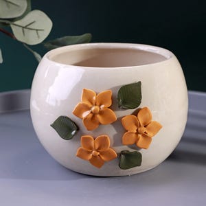 May include: A cream-coloured ceramic planter with a rounded shape. It features three-dimensional orange flowers and green leaves. The glossy finish adds a touch of elegance to this decorative piece.