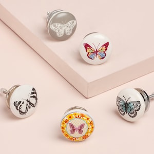 Peut inclure: Cinq boutons de meubles décoratifs avec des motifs de papillons. Les boutons présentent une variété de couleurs, dont le gris, le rouge, le jaune et le bleu. Les boutons sont ronds avec une base en métal et sont posés sur un fond rose.