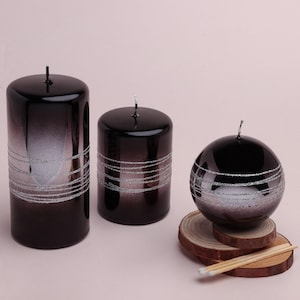 May include: Three decorative candles in varying sizes. The candles are black with a gradient effect and silver glitter bands. One candle is cylindrical, one is shorter and cylindrical, and the third is spherical. The candles sit on wooden slices.