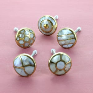 May include: Five decorative cabinet knobs with brass rims and various inlaid designs. The knobs feature white and iridescent shell patterns, including geometric and striped designs. Each knob has a silver screw for attachment.