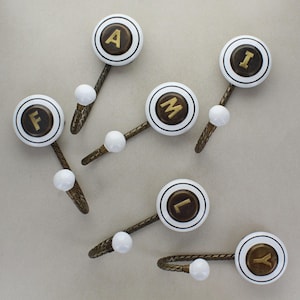 May include: A collection of decorative wall hooks. Each hook has a white circular knob with a gold letter in the centre, including A, F, I, L, M, and Y. The hooks have a bronze-coloured metal stem and a small white ball. The hooks are suitable for any room.