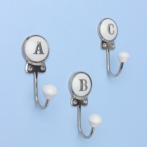 Può includere: Tre ganci da parete decorativi con finitura argentata e pomelli in ceramica bianca. Ogni gancio ha una parte superiore circolare in ceramica bianca con una lettera nera: A, B o C. I ganci sono montati su una parete azzurra.