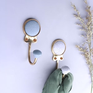 May include: Two wall hooks featuring a gold-toned metal frame with a circular top inlay of light blue ceramic with a crackle finish. Each hook includes two small amber-colored decorative accents and a rounded light blue ceramic tip.