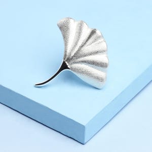 Op de afbeelding: Een zilverkleurige ginkgo blad broche. De broche is een gedetailleerde replica van een ginkgo blad, met getextureerde aderen en een gebogen steel. Het blad is geplaatst op een lichtblauwe ondergrond.