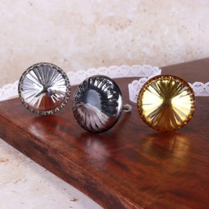 May include: Three round cabinet knobs with a fluted, sunburst-like design. The left knob is polished silver, the centre knob is dark metallic grey, and the right knob is a warm, reflective gold tone. Each knob features a central mounting post.