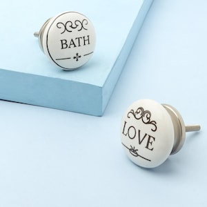 Könnte beinhalten: Zwei weiße Keramik-Schrankknöpfe mit schwarzem Text und dekorativen Verzierungen. Auf einem Knopf steht "BATH" und auf dem anderen "LOVE". Beide Knöpfe haben einen silberfarbenen Metallsockel und eine Schraube.
