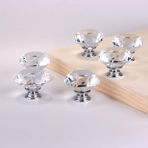 May include: A collection of five clear crystal cabinet knobs with a diamond-like faceted design. Each knob has a polished silver-tone base. The knobs are arranged on a light-colored wooden surface, showcasing their reflective qualities.