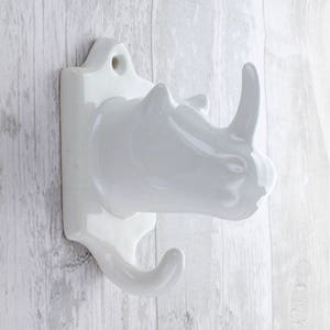 May include: White ceramic rhinoceros wall hook with two hooks. The hook is shaped like a rhinoceros head with a raised horn.