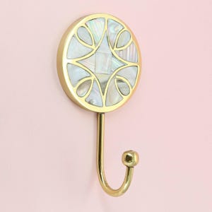 Op de afbeelding: Een decoratieve wandhaak met een cirkelvormig ontwerp. De haak heeft een goudkleurige metalen afwerking en een ronde voorkant ingelegd met iriserend parelmoer. De haak zelf buigt naar beneden en eindigt in een kleine bal. De haak is gemonteerd op een roze muur.