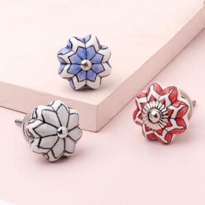 Peut inclure: Trois boutons de meuble en céramique avec des motifs floraux. L'un est blanc et gris, un autre est rouge et blanc, et le troisième est bleu et blanc. Chaque bouton a une base et une vis en métal argenté.