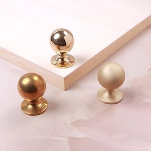 Puede incluir: Tres pomos esféricos para armarios con bases circulares. Un pomo tiene un acabado dorado pulido, otro un acabado de latón cepillado y el tercero un acabado dorado mate. Los pomos están dispuestos sobre y frente a una tabla de madera clara.