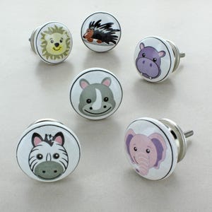 May include: A set of ceramic cabinet knobs with animal designs: lion, zebra, elephant, rhino, hippo, and hedgehog. Each knob has a white background, black border, and a silver metal base. Ideal for children's furniture.