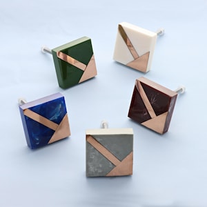 Op de afbeelding: Vijf vierkante kastknoppen met een geometrisch ontwerp. De knoppen zijn gemaakt van verschillende gekleurde materialen, waaronder groen, wit, blauw, grijs en rood. Elke knop heeft een gouden metalen accent.