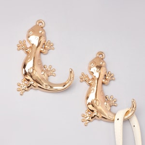 May include: Two gold-colored lizard charms with a shiny finish. The charms have detailed legs, a curved tail, and a loop at the top for hanging. One charm is attached to a white strap. The lizards have a textured surface.