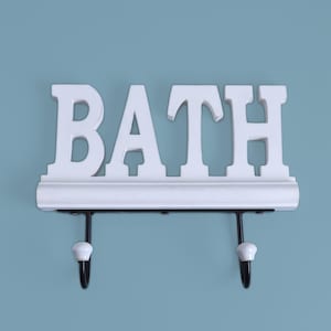 May include: White wooden wall decor with the word "BATH" in large capital letters. The sign has two black hooks with white ceramic knobs. The sign is mounted on a light blue wall.