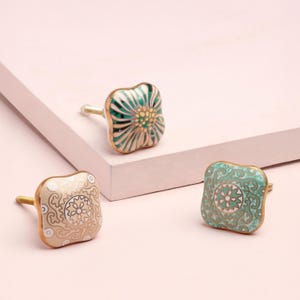 May include: Three decorative cabinet knobs with brass stems. One knob is square with a cream background and a gold floral design. Another is square with a teal background and a gold floral design. The third knob is square with a green and white floral design.