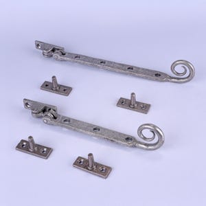 May include: Two antique-style window stays with matching mounting plates. The stays are a distressed silver colour, with a decorative scroll end. The mounting plates are rectangular with a single post. Hardware for windows or cabinets.