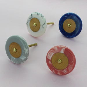 Peut inclure: Quatre boutons de meuble décoratifs de différentes couleurs : blanc avec marbrure grise, bleu, rouge avec marbrure blanche et gris avec marbrure blanche. Chaque bouton a un centre en bois et une vis en laiton.