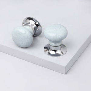 May include: Two cabinet knobs with a crackle glaze finish. The knobs are light blue and have a round shape. They are mounted on a shiny, silver-colored base. The knobs are designed for use on cabinets or drawers.