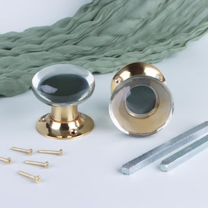 May include: Two round door knobs featuring clear glass tops and polished brass bases. The set includes four small gold-toned mounting screws and two square metal spindles for installation, presented as a complete hardware assembly.
