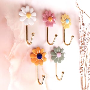 May include: A set of five wall hooks featuring flower-shaped heads in white, pink, yellow, orange, and sage green. Each hook has a gold-toned metal J-shaped arm with a small ball tip at the end. The flower centers are yellow, blue, or pink.