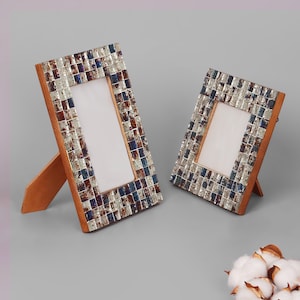 Pode incluir: Duas molduras retangulares com bordas de mosaico em tons de prata, azul e marrom. As molduras têm costas e suportes de madeira e são anguladas para o espectador. As molduras estão vazias, prontas para fotos.