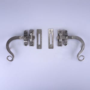 May include: Two antique-style, silver-colored window latches. Each latch has a curved handle with a decorative scroll end. The set includes two rectangular strike plates and two latch mechanisms, all with a rustic, hammered finish.