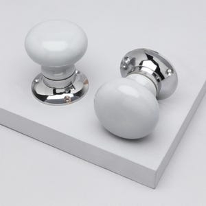 May include: Two white ceramic door knobs with a polished chrome base. One knob is upright, and the other is lying on its side. The knobs have a rounded shape and are set against a white background.