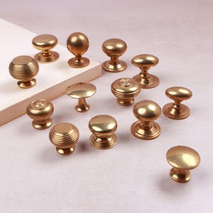 May include: A collection of thirteen brass cabinet knobs in various shapes and sizes. Some knobs feature a smooth, rounded surface, while others display a ribbed or concentric circular pattern. Each knob has a circular base and a central stem.