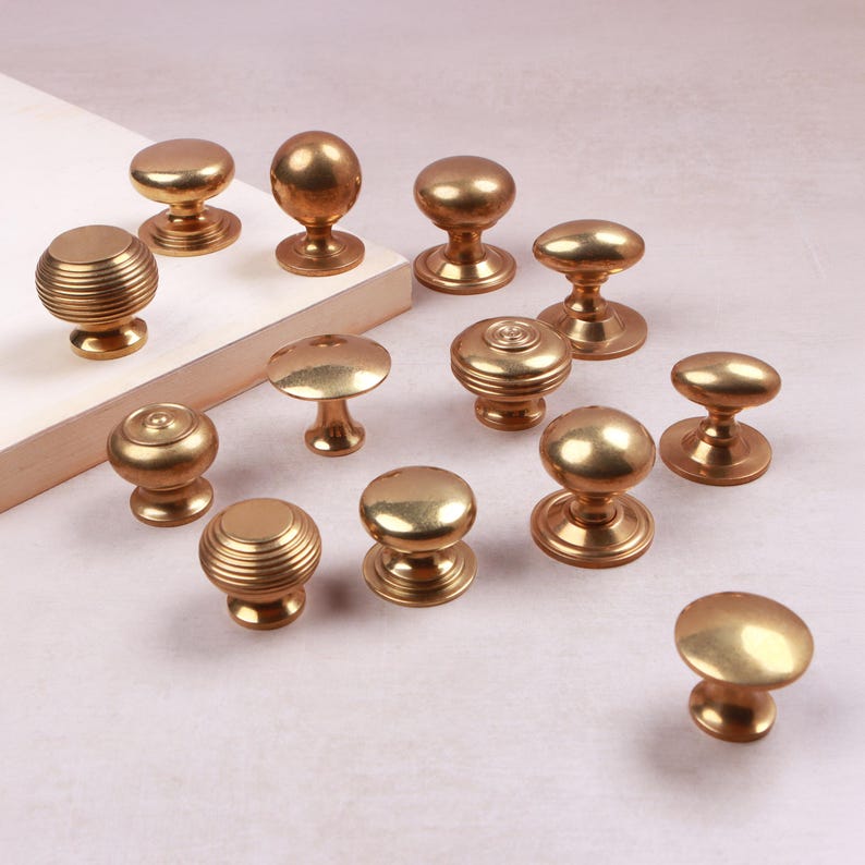 May include: A collection of thirteen brass cabinet knobs in various shapes and sizes. Some knobs feature a smooth, rounded surface, while others display a ribbed or concentric circular pattern. Each knob has a circular base and a central stem.