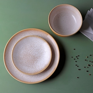 Può includere: Un set di stoviglie in ceramica, composto da due piatti e una ciotola. I piatti sono impilati, con un piatto più grande sotto uno più piccolo. La ciotola è rotonda. Tutti i pezzi hanno un design bianco e marrone maculato e uno sfondo verde.
