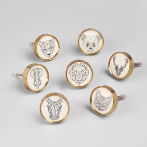 May include: Set of 7 gold cabinet knobs with white ceramic inserts featuring geometric animal designs. The animals include a lion, a bear, a deer, an elephant, a zebra, a cat, and a panther.