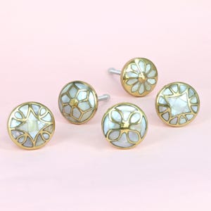 May include: Five decorative cabinet knobs with gold-colored metal bases and white mother-of-pearl inlay. The knobs feature various geometric and floral designs. The knobs are arranged on a light pink background.