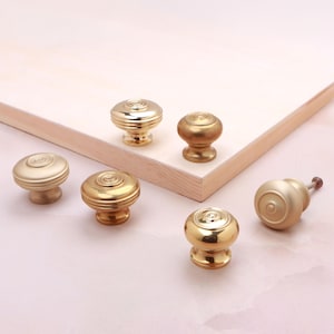 Peut inclure: Un ensemble de six boutons de placard ronds dans diverses finitions dorées, incluant des textures polies et brossées. Chaque bouton présente un design circulaire à gradins avec un point central, affichant un éclat métallique et une forme robuste.