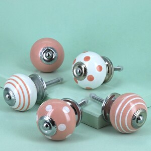 Può includere: Una collezione di pomelli decorativi per mobili. I pomelli hanno una forma rotonda con base e vite in metallo. I disegni includono pois rosa e bianchi, strisce rosa e bianche e rosa tinta unita. Misure in centimetri.