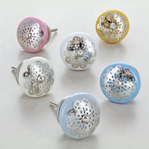 Puede incluir: Seis pomos decorativos para armarios en varios colores: rosa, blanco, amarillo y azul. Cada pomo presenta un diseño de filigrana plateada sobre una base de color sólido. Los pomos son redondos y tienen un tornillo para una fácil instalación.
