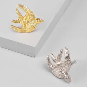 Puede incluir: Dos tiradores decorativos con forma de pájaro, uno dorado y otro plateado. Los tiradores presentan detalles texturizados y son adecuados para muebles. Ideales para armarios o cajones.