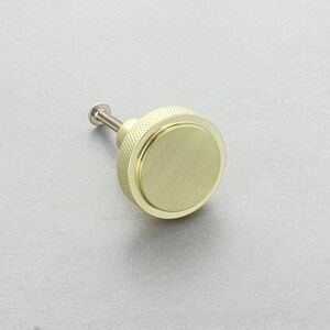 Könnte beinhalten: Ein goldfarbener Schrankknopf mit einer kreisförmigen, flachen Oberfläche und einem strukturierten, zylindrischen Griff. Der Knopf hat einen kleinen, silberfarbenen Stiel zur Befestigung. Das Design ist schlicht und modern.