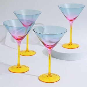 Może przedstawiać: Cztery kieliszki do martini z gradientem od niebieskiego do różowego w czaszy, żółtymi nóżkami i podstawami. Kieliszki są ułożone na białej powierzchni, prezentując ich kolorowy design. Idealne do koktajli.