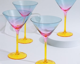 May include: Four martini glasses with a gradient of blue to pink in the bowl, yellow stems and bases. The glasses are arranged on a white surface, showcasing their colorful design. The glasses are ideal for cocktails.