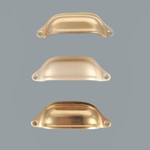 Peut inclure: Trois poignées de tiroir en forme de coquille en métal disposées verticalement. Chaque poignée présente une forme incurvée lisse avec deux trous de vis circulaires sur les rebords latéraux. Finitions or poli, or mat et laiton antique.