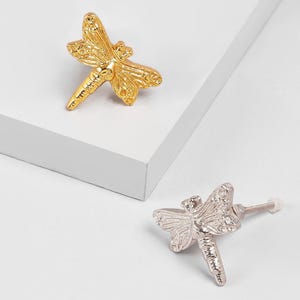 May include: Two decorative dragonfly cabinet knobs. One is gold-colored, and the other is silver-colored. The knobs are made of metal and have detailed wing and body designs. The knobs are designed to be attached to a cabinet or drawer.