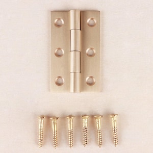May include: A rectangular brass butt hinge with a brushed finish featuring three countersunk screw holes on each leaf. Below the hinge are six matching brass wood screws with flat heads, arranged in a horizontal row for hardware installation.