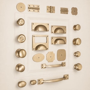 May include: A collection of brass-toned cabinet hardware including various round knobs, cup pulls, rectangular label holders, hinges, and long drawer handles. The hardware features a matte finish and is arranged in a grid-like display pattern.